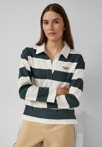 Jonge vrouw met lang bruin haar, gekleed in een groen-wit gestreept rugbyshirt met een kraag en beige broek, staande met de armen over elkaar.