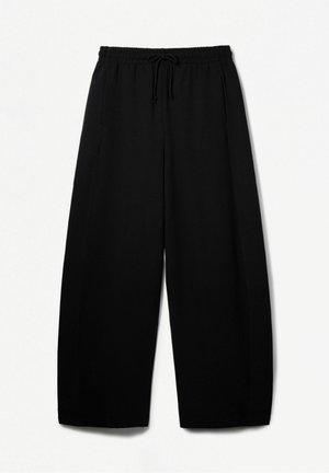 Femme portant un sweat-shirt noir oversize, un pantalon noir à jambes larges, des baskets blanches, les mains sur les hanches devant un fond blanc uni.