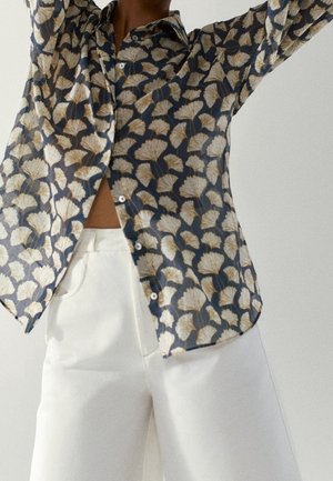 Personne portant une chemise bleu foncé à boutons avec des motifs de feuilles blanches et un pantalon blanc taille haute avec des poches, bras levés au-dessus de la tête.