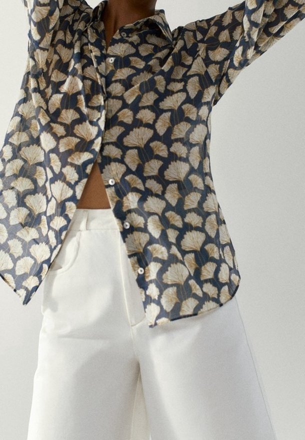 Personne portant une chemise bleu foncé à boutons avec des motifs de feuilles blanches et un pantalon blanc taille haute avec des poches, bras levés au-dessus de la tête.