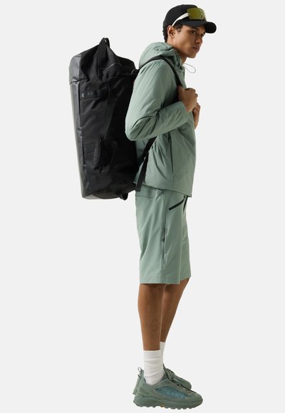 Young man in outdoor gear wearing a large black backpack, green jacket, matching shorts, cap, and sunglasses on head.