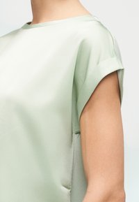 Κοντινό πλάνο ενός ατόμου που φοράει ένα κοντομάνικο ανοιχτοπράσινο σατέν μπλουζάκι με διπλωμένο μανσέτι στο μανίκι.