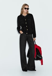 Donna che indossa un cardigan nero a trama con bottoni dorati, jeans neri a gamba larga, occhiali da sole neri, mentre tiene una borsa nera con un maglione rosso.
