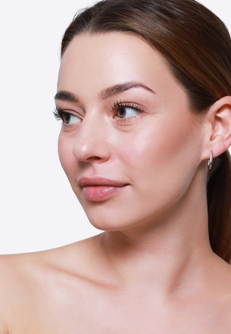 Primo piano di un volto con pelle chiara, texture liscia e trucco leggero. Le caratteristiche includono occhi marroni, sopracciglia definite e orecchini a cerchio.