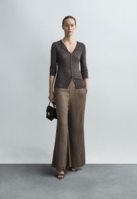 Brūns garroku kardigans ar pogām priekšpusē, kopā ar plašām brūnām biksēm. Vienā rokā tur mazu melnu rokassomiņu.