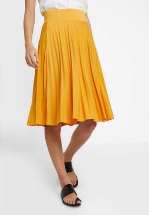 Kvinde iført et knælængde, plisseret gult nederdel med bred linning, hvid top og sorte åbne tå-sandaler, stående på en hvid baggrund.