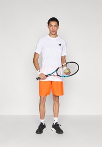 Wit atletisch shirt met korte mouwen en zwart logo, oranje korte broek, zwarte sneakers, polsbandjes; houdt een tennisracket vast met een bal.