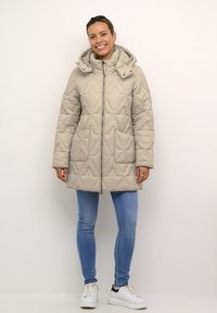 Gaišbeige quilted jaka ar kapuci, ar dimanta rakstiem, divām priekšējām kabatām un priekša rāvējslēdzēju. Nēsāta ar gaiši zilām džinsiem un baltām sporta kurpēm.