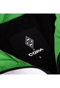 Groene jasinterieur met zwarte netvoering. Beschikt over een zwart etiket met witte tekst, logo en maat "S." Ritsrand zichtbaar.