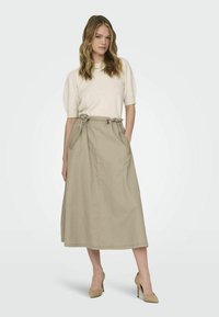 Beige sukně po kolena z lehkého materiálu s páskem na stažení a bočními kapsami. V kombinaci s krémovým topem s krátkým rukávem a béžovými podpatky.