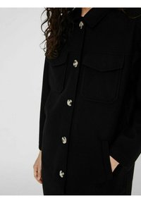 La prenda exterior negra presenta un diseño de abotonado, dos bolsillos en el pecho, botones plateados y una textura suave. Incluye mangas largas y bolsillos laterales.
