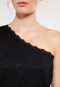 Haut en dentelle noir présentant un design asymétrique à une épaule avec des motifs floraux complexes et une finition texturée, mettant en avant des détails élégants.