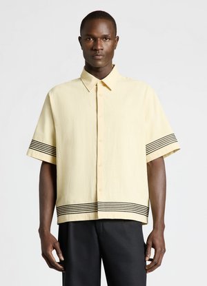 Homme portant une chemise jaune clair à manches courtes avec des rayures noires horizontales sur les manches et l'ourlet, associée à un pantalon noir.