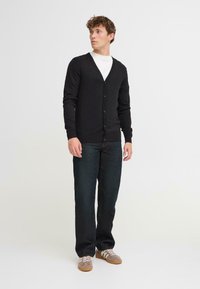 Svart cardigan-genser med V-hals, knappelukking og ribbestrikkede mansjetter, båret over en hvit skjorte sammen med mørkeblå jeans og grå sneakers.