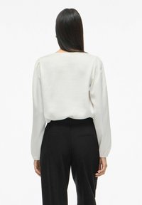 Weiße Bluse mit langen Ärmeln, leicht gepufften Schultern und glatter Textur. Kombiniert mit schwarzen, maßgeschneiderten Hosen.