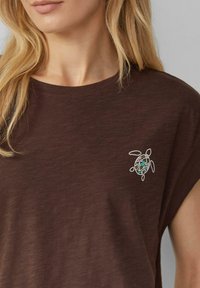 Blonde woman wearing a brown shirt with a small colorful turtle graphic on the chest near the collar.