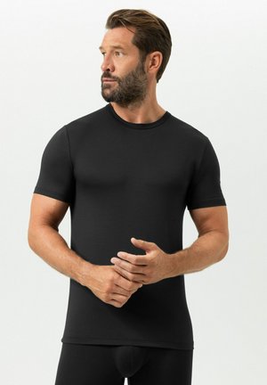 Man met baard die een zwarte, nauwsluitende korte mouw shirt draagt, kijkt naar links, met handen op de taille gevouwen tegen een effen witte achtergrond.