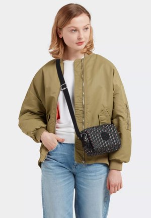 Giovane donna che indossa una giacca bomber marrone chiaro, una camicia bianca, jeans blu e una borsa a tracolla con un motivo e una tracolla nera.