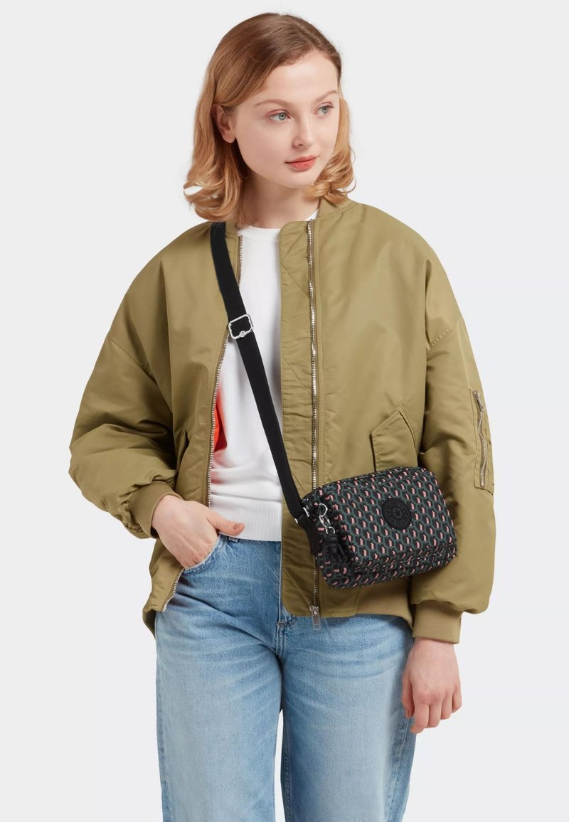 Giovane donna che indossa una giacca bomber marrone chiaro, una camicia bianca, jeans blu e una borsa a tracolla con un motivo e una tracolla nera.