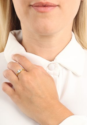 Anillo de oro con una banda lisa que presenta una piedra central de corte redondo y transparente, llevado en una mano contra una camisa blanca de cuello.