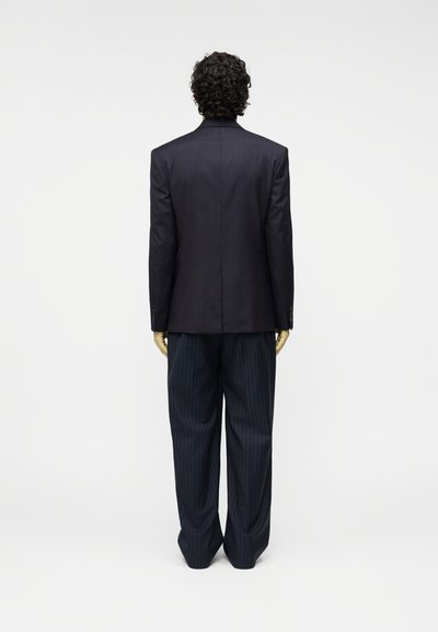 Σακάκι σε κομψή γραμμή σκούρου μπλε ναυτικού με κομψούς γιακάδες με εγκοπές, συνδυασμένο με παντελόνια φαρδιά με κάθετες ρίγες. Λείο ύφασμα, καθαρές γραμμές, δομημένη σιλουέτα.