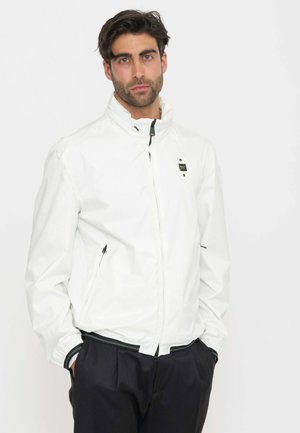 Uomo con capelli scuri che indossa una giacca bianca con zip e tasche con zip e pantaloni neri, in piedi davanti a uno sfondo semplice.