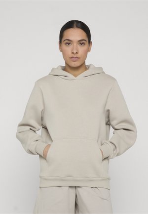 Junge Frau mit dunklen Haaren, die einen beigen Kapuzenpullover und passende Hose trägt, steht mit den Händen in den Taschen des Kapuzenpullovers vor einem einfarbigen Hintergrund.