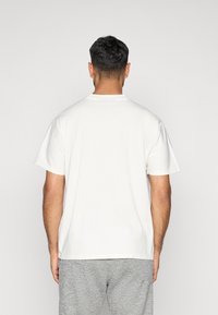 T-shirt de algodão branco com mangas curtas, decote redondo, vista traseira lisa contra um fundo neutro, usada com calças de moletom cinzas.