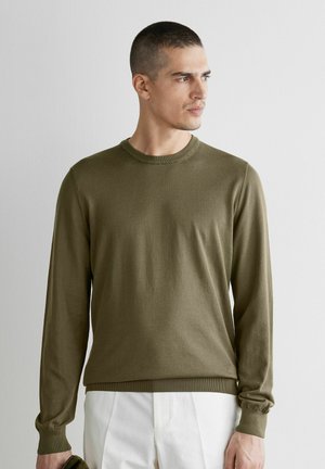 Homme portant un pull à manches longues vert olive et un pantalon blanc, regardant sur le côté contre un fond clair uni.
