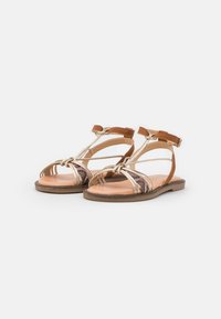 Tan vlakke sandalen met gouden banden, met een geweven ontwerp en open teen. Verstelbare enkelband voor een veilige pasvorm. Vlakke zool met minimale profiel.