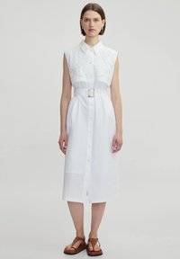 Witte mouwloze jurk met een kraaghalslijn, complexe borduursels op het lijfje en een ceintuur in de taille, met een knoopsluiting aan de voorkant en een split.