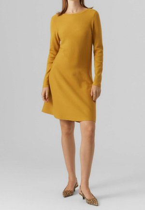 Femme portant une robe jaune moutarde à manches longues associée à des chaussures slingback à talons bas imprimées léopard, debout devant un fond uni.