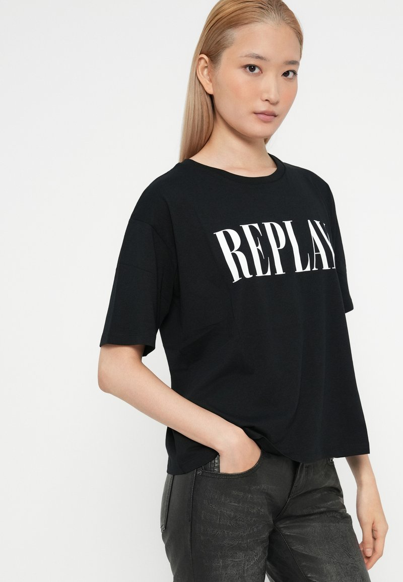 Camiseta de algodón negra con mangas cortas y cuello redondo, que presenta un gran logo blanco de "REPLAY" en la parte frontal. Corte casual.