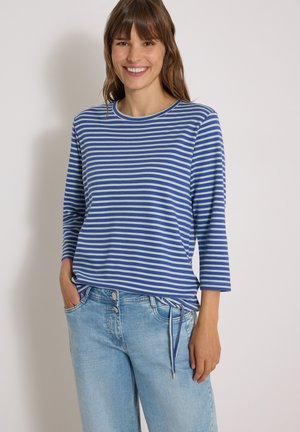 Femme souriante portant un t-shirt à manches longues bleu et blanc à rayures et un jean bleu clair, les mains dans les poches.