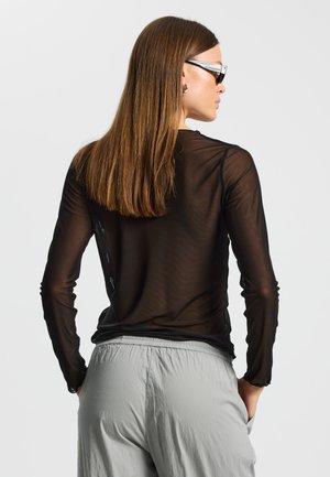 Czarna przezroczysta bluzka z długim rękawem o dopasowanym kroju, nałożona na jasnoszare, luźne spodnie. Gładka faktura, subtelny połysk.