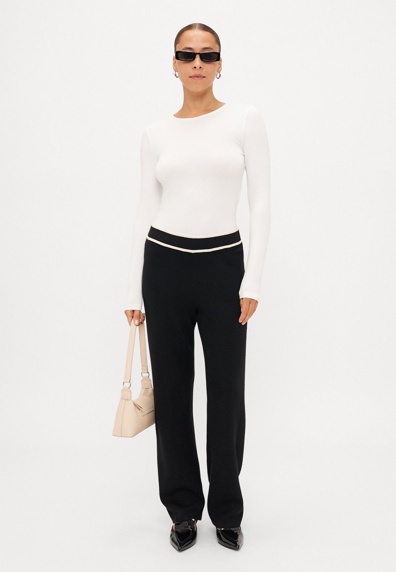 Un top bianco a maniche lunghe aderente abbinato a pantaloni neri a gamba larga a vita alta, scarpe a punta nere e una borsa beige chiaro.