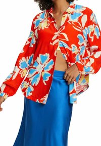 Blouse rouge avec un motif floral bleu et jaune. Dotée d'une fermeture à boutons, de manches longues et d'une coupe décontractée. Associée à une jupe en satin bleu.