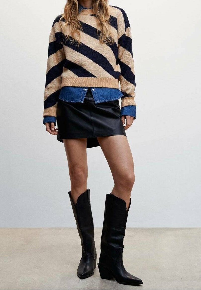 Femme portant un pull beige et noir à rayures diagonales par-dessus une chemise bleue, une mini-jupe en cuir noire, et des bottes noires montantes, debout sur un sol gris.