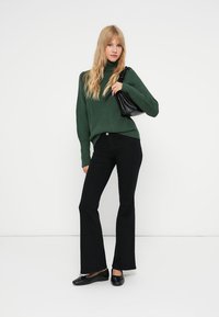 Suéter verde de malha com gola alta, jeans pretos flare, sapatos pretos rasos e uma pequena mala preta. Modelo posando contra um fundo branco.