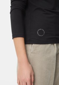 Czarny top z długim rękawem o dopasowanym kroju, z okrągłym napisem z logo u dołu, zestawiony z jasnobrązowymi spodniami sztruksowymi.