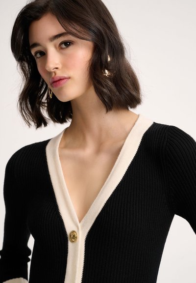 Cardigan noir à côtes avec un col en V crème, fermeture à un seul bouton doré et manches longues ajustées, mettant en valeur un tissu tricoté texturé.