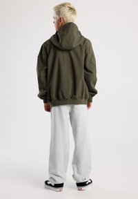Olive green hoodie with a lined hood, loose fit, and ribbed hem, paired with light gray sweatpants. Black and white sneakers complete the look.