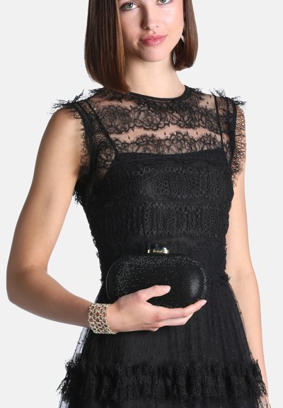 Femme portant une robe en dentelle noire, tenant une petite pochette noire scintillante et portant un bracelet en perles et or à son poignet.