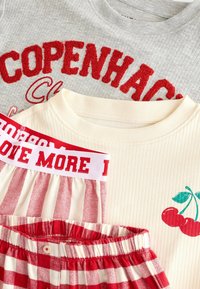 Drei lässige Oberteile sind gestapelt: ein graues Rippoberteil mit rotem "COPENHAGEN"-Text, ein cremefarbenes Rippoberteil mit einem Kirschdesign und rot karierte Shorts mit einem roten Bund.