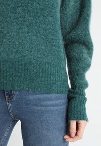 Turkoosi villaneule, jossa on ribbireunat ja hihansuut, pehmeällä tekstuurilla. Yhdistetty sinisiin farkkuihin, jotka esittelevät osan asusta.