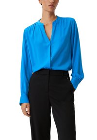Blaue Seidenbluse mit langen Ärmeln, einem Mandarin-Kragen und einer lockeren Passform. Getragen mit schwarzen Hosen, die ein schlichtes Design und Taschen haben.