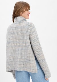 Vrouw die glimlacht, gekleed in een losse, geribbelde lichtgrijze turtleneck trui met subtiele blauwe en beige strepen, gecombineerd met lichtblauwe jeans.