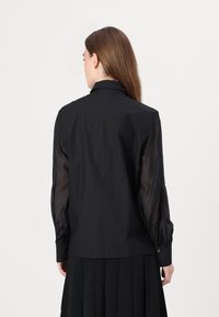 Maglietta nera con corpo in tessuto solido e maniche a sbuffo trasparenti. Design con colletto e polsini con bottone, creando un effetto visivo a strati.