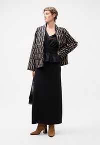 Veste à motifs noir et beige sur un top peplum noir et une longue jupe noire. Bottes à talons beiges. Le mannequin tient un sac noir.