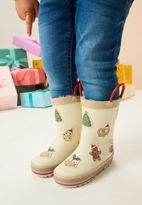 Weiße Gummiregenstiefel mit festlichen Feiertagsmustern, darunter Kekse und Bäume. Sie haben rote Griffe und eine mehrfarbige Sohle.